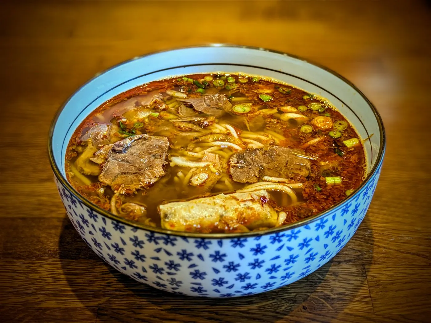 Bowl of bún bò Huế at Fire Pho in Huntington Beach, CA — chili-red lemongrass broth with thick round rice noodles, sliced beef shank, beef tendon, and chả lụa, topped with green onion.